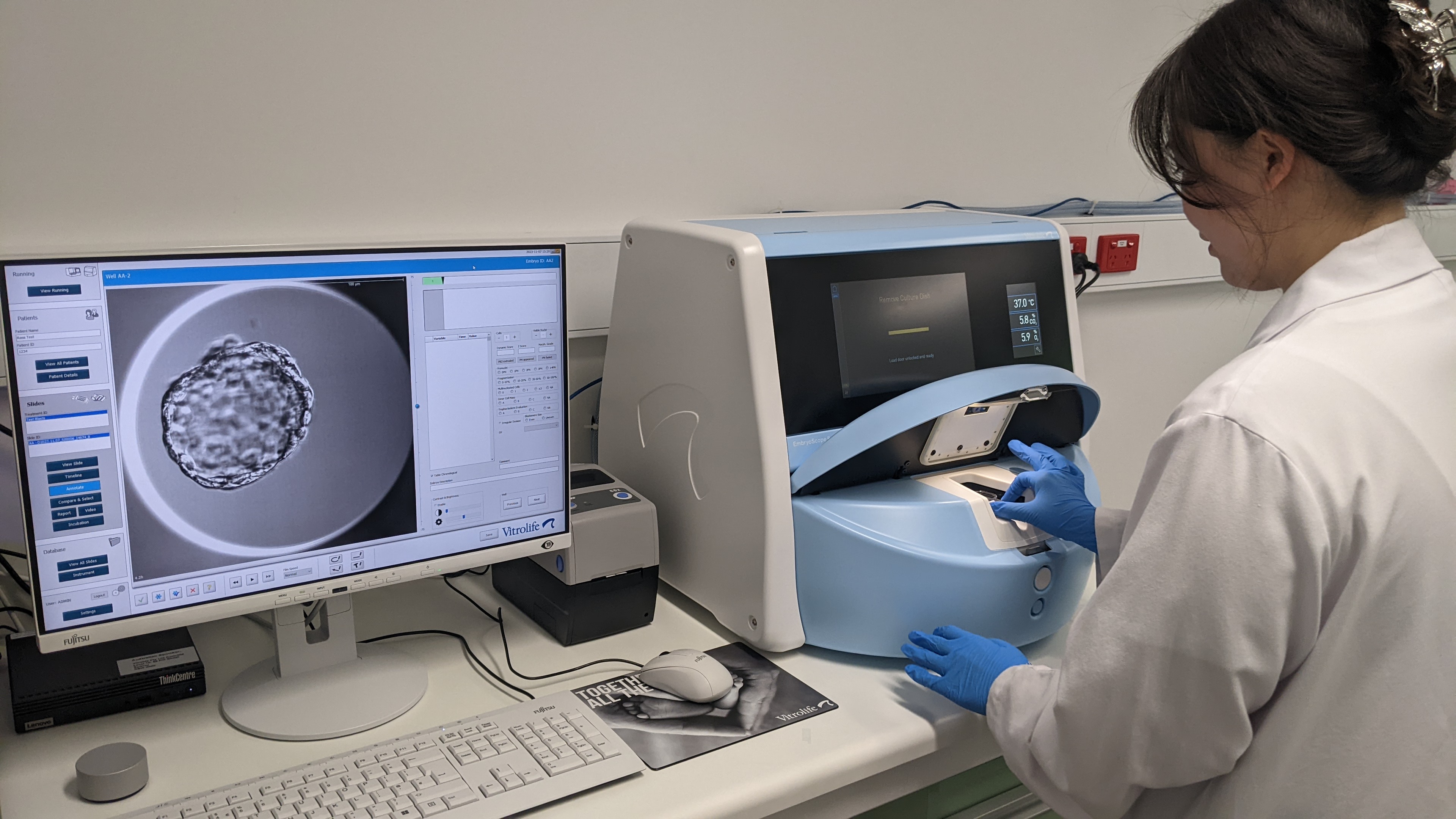 A researcher wearing a white lab coat and blue rubber gloves is using a light blue machine on the right hand side of the photo. On the left hand side an image from the EmbryoScope is being displayed on a computer screen. The image displayed on the computer screen is of a mouse embryo