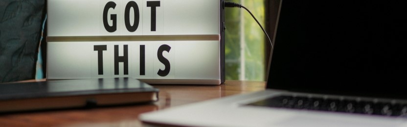 An open laptop is on a table next to a notebook. In the background is a LED lamp sign with the words &ldquo;You Got This&rdquo;.