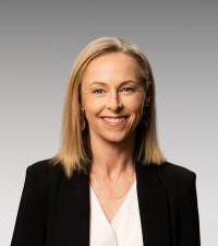 A person with long blond hair wearing a black blazer and white shirt smiling at the camera.
