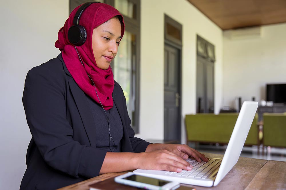 A teacher with a head scarf and headphones working on laptop