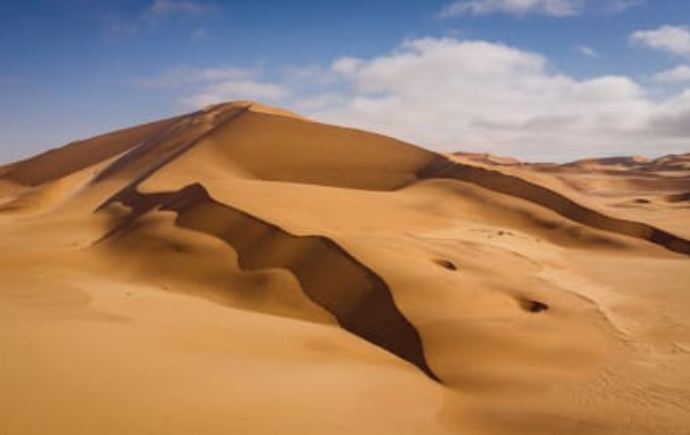A wide landscape photograph of rolling golden-yellow sand dunes in a desert. Sharp ridges across the dunes create strong dark shadows on one side and sunlit slopes on the other. In the background, a light blue sky is dotted with soft, scattered