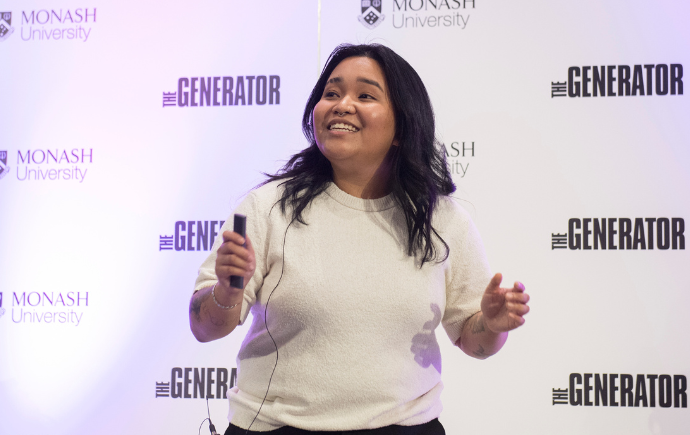 A woman entrepreneur pitching on stage in front of a screen with the Monash and Generator logos