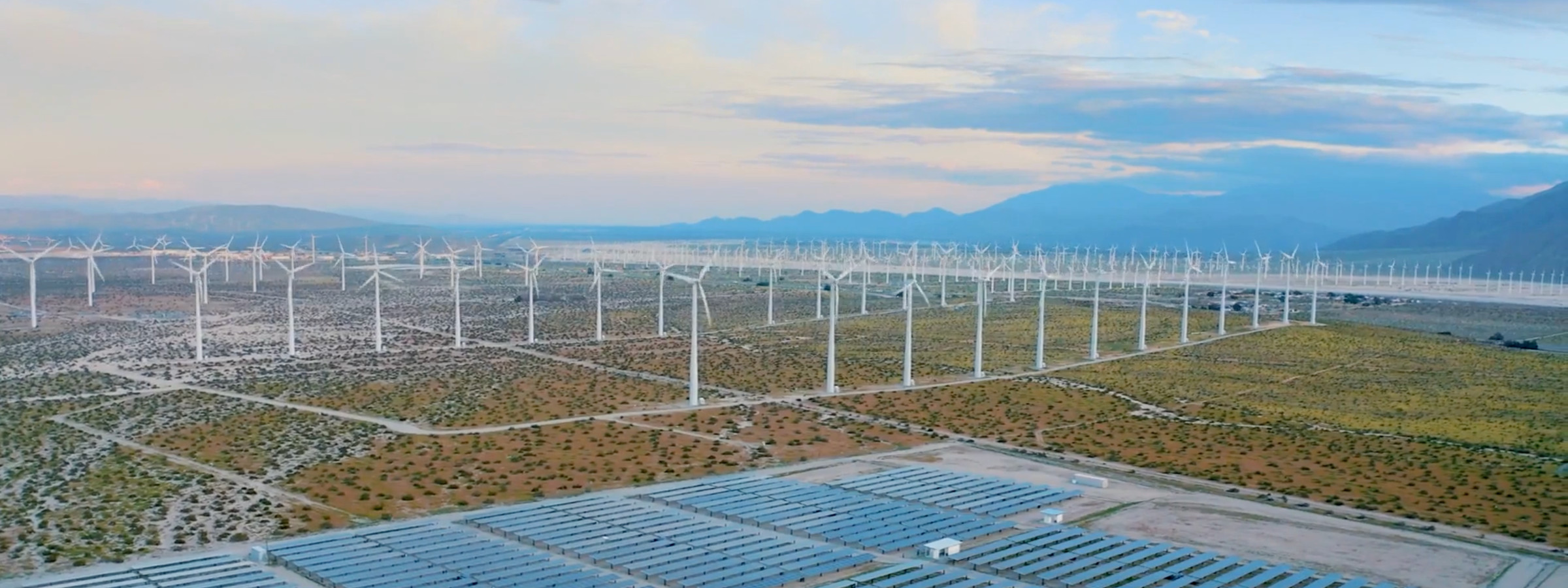 Aerial view of a flat shrub plain with a solar farm in the foreground, a wind farm in the midground and low hills and mountains in the distance