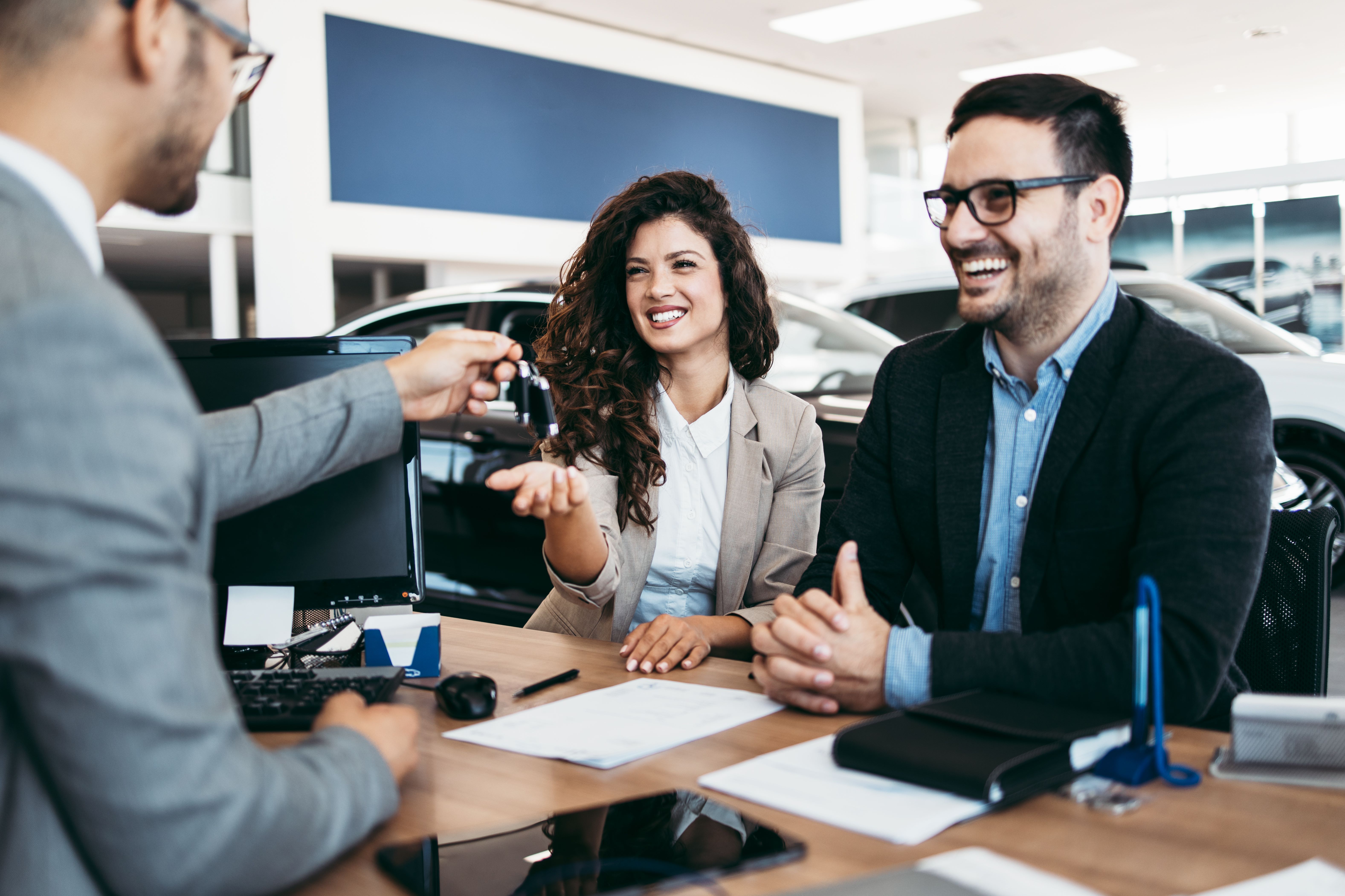 A smiling woman and man sitting across the desk from another man in a suit as he hands some car keys to the woman