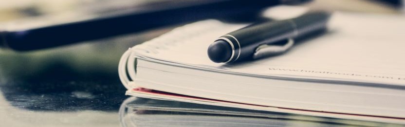 Close up of a black biro pen and a lined diary-style notebook.
