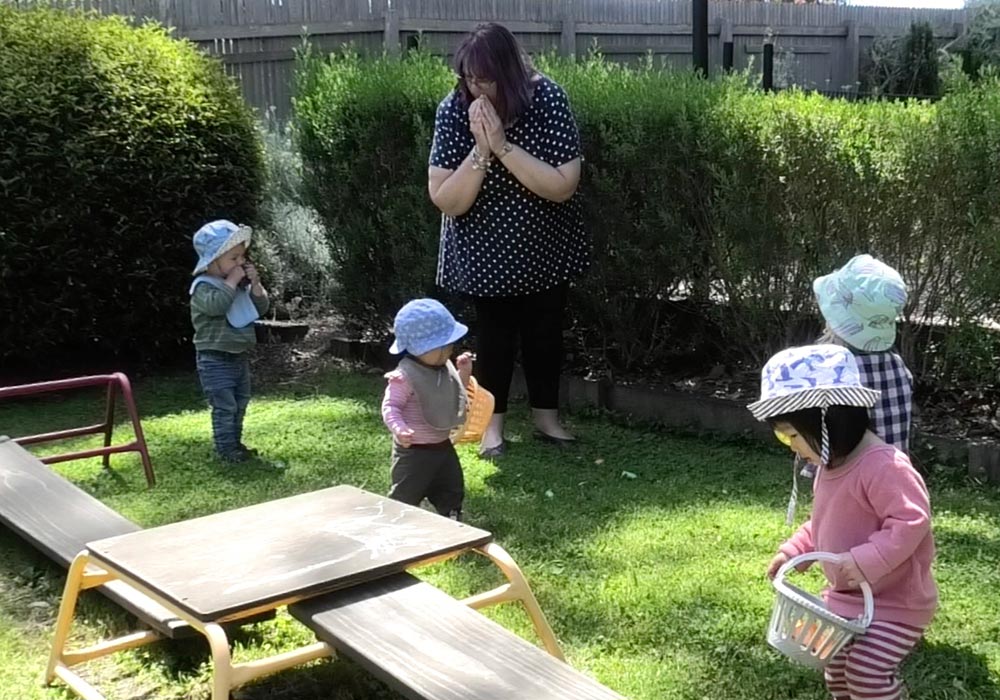 Four toddlers in the backyard at kinder, searching for things to put into their baskets, and teacher playing along.