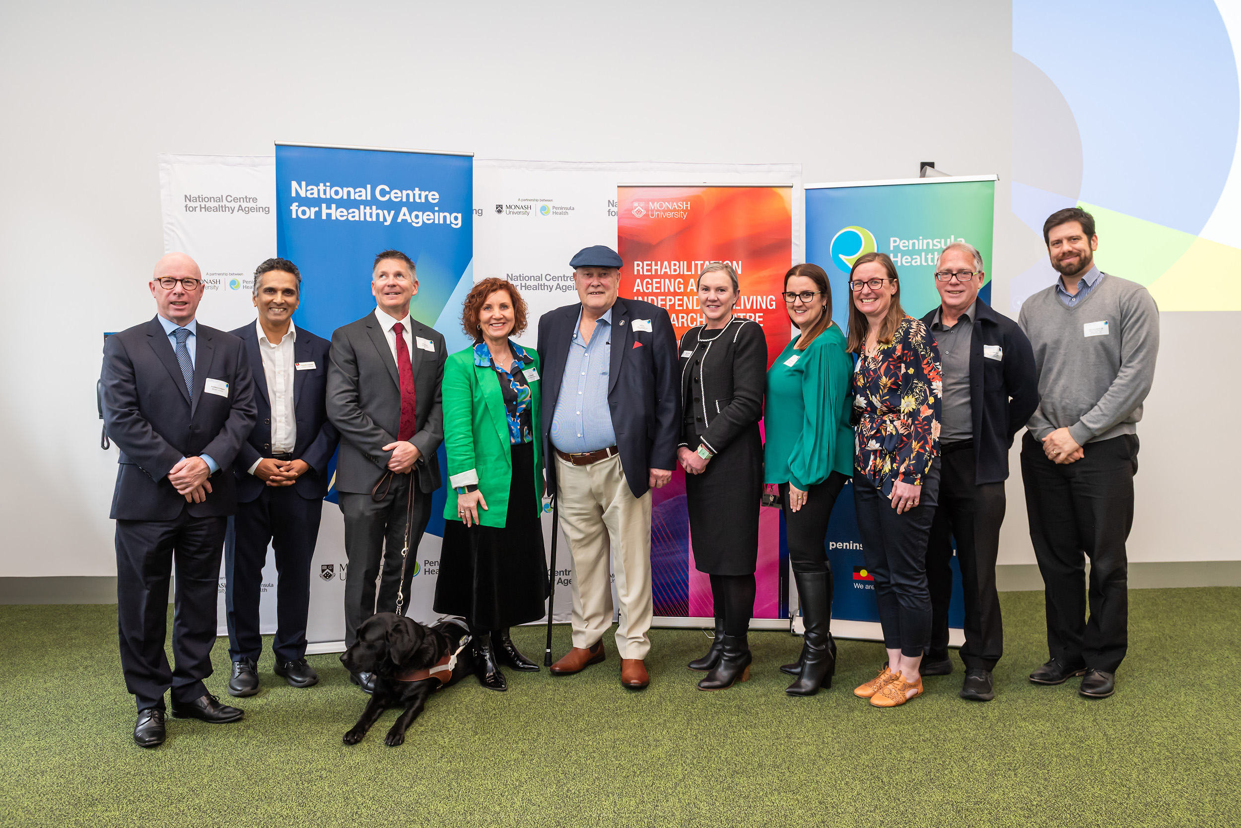 A photo of 10 people standing in front of three organisation banners (National Centre for Healthy Ageing; Rehabilitation, Ageing and Independent Living (RAIL) Research Centre; Peninsula Health) smiling at the camera. A black Labrador guide dog is lying in front of a person.