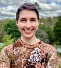 A headshot of Jacinta smiling at the camera. She has short brown hair and wears a brown floral shirt.