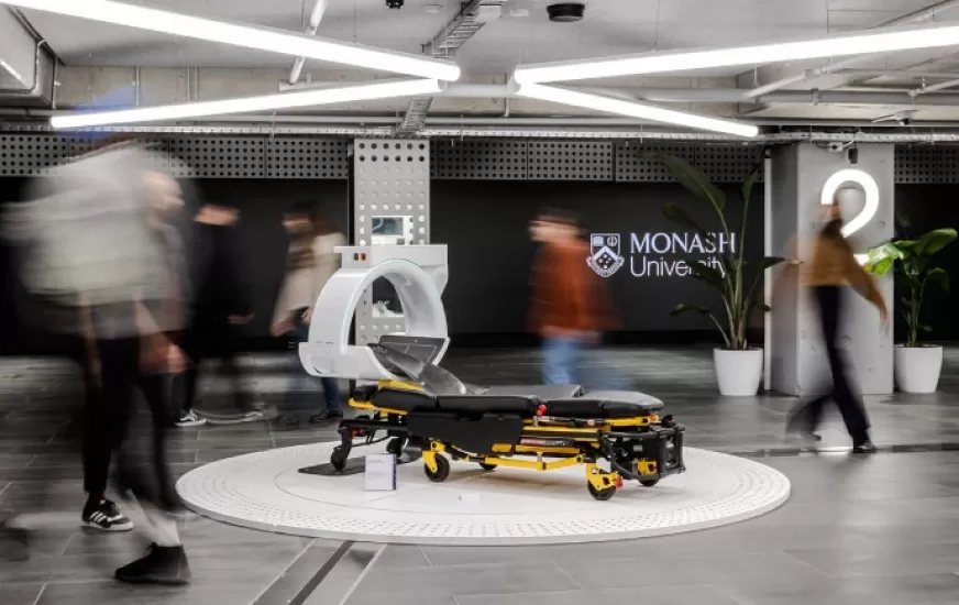 People walk about an advanced CT scanning trolley in the centre of a large room, the Monash logo can be seen on the far wall, prominently.