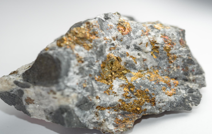 A rough gray rock sample with irregular surfaces, containing shiny gold-colored metallic flecks and veins, photographed against a plain light background.