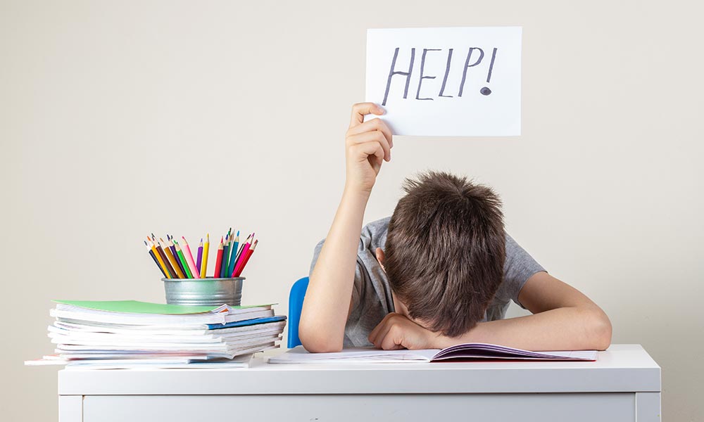 a boy sitting at a school desk with a sign above his head that says "help"