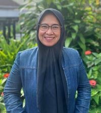A person standing outside, wearing glasses, a head scarf and a jeans jacket smiling at the camera.