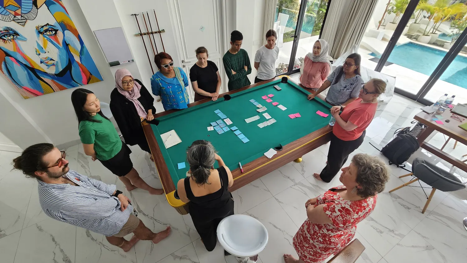 A photo from above of twelve Indonesian and Australian researchers standing in bare feet around a billiard table, listening to one of the researchers speak. In the background is a swimming pool, vivid artwork of a woman and a table with papers, water bottles and reading glasses. There are bags on the floor. On the billiards table are approx 20 post-it notes of different colours with text on them, arranged in clear patterns.&nbsp;&nbsp;All researchers have their heads and bodies turned towards one researcher in black with her hair in a ponytail, who is gesticulating as she speaks.