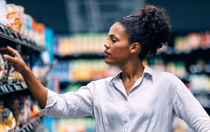 A person stands in a grocery store aisle, reaching toward items on a shelf. The shelves on both sides are stocked with packaged food products, and the background is softly blurred, emphasising the action of selecting an item.