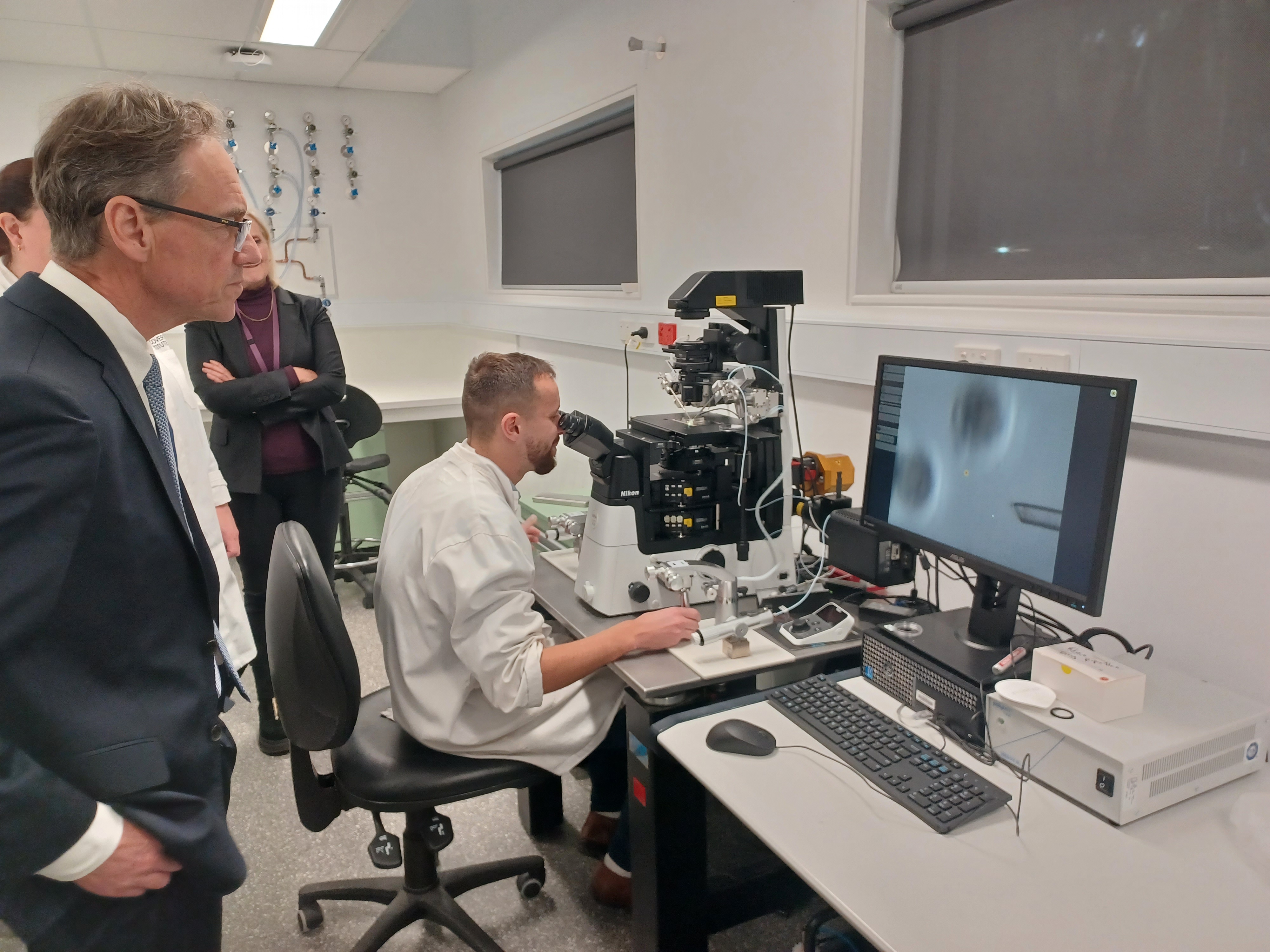 A mitoHOPE researcher is seated at a laboratory workstation, operating an advanced microscope to perform mitochondrial donation. Greg Hunt stands nearby, partially facing away from the camera, and watching the researcher working. The workstation includes various scientific instruments and a computer monitor displaying an enlarged image of the mouse eggs. The room has white walls and there’s a window with blinds drawn down.