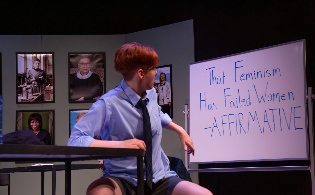 An actor playing a school boy sits at a desk, turning to face the whiteboard behind them that has the words 
