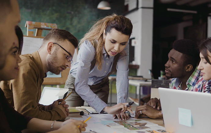 A group of people sitting at a desk, huddled over a document with one person standing and pointing to the document on the desk.